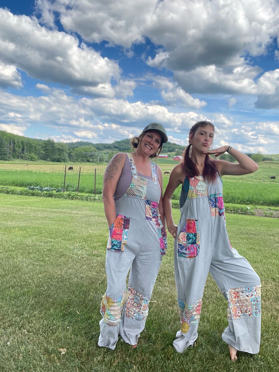 Two women smiling and posing outdoors in a green farm field, wearing gray Patchwork Boho Overalls with colorful patchwork.