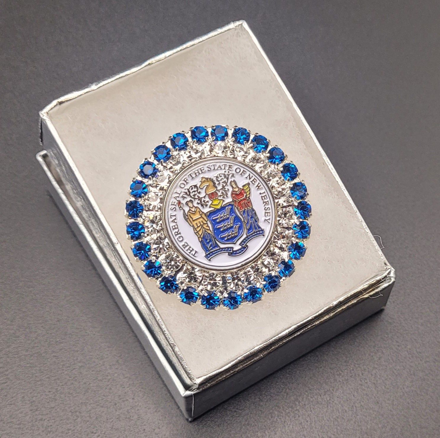 A jeweled brooch featuring the Great Seal of the State of New Jersey with blue jewels around the rim in a white box.