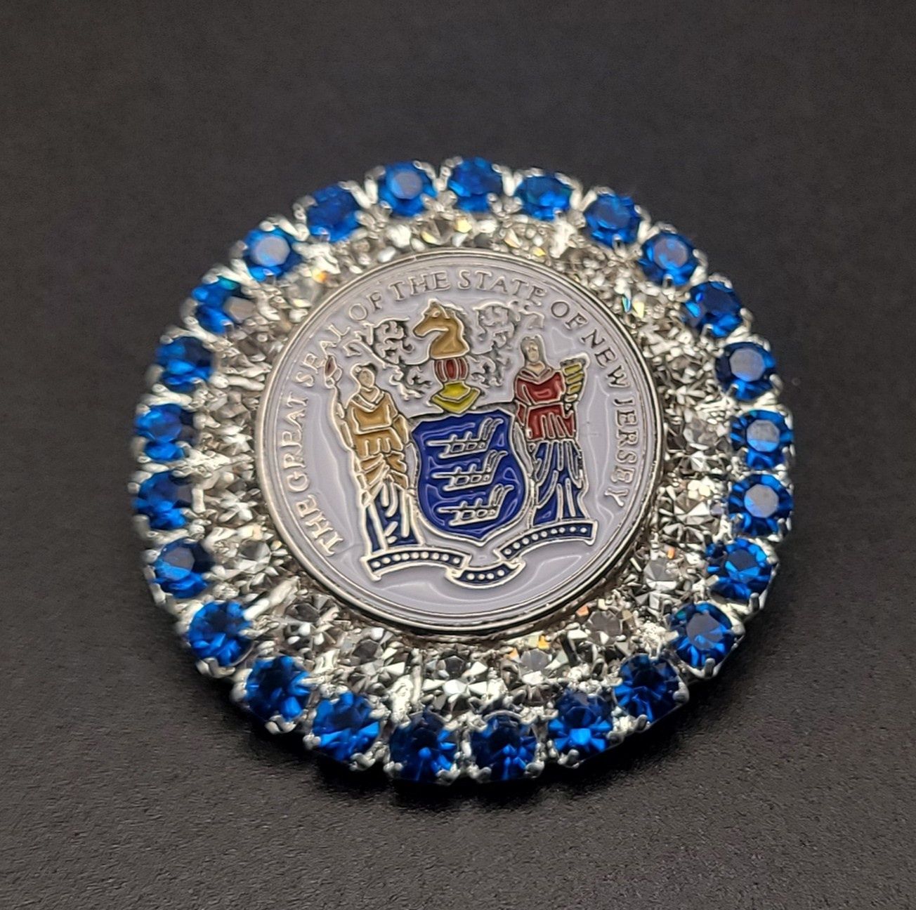 A jeweled brooch featuring the Great Seal of the State of New Jersey with blue jewels around the rim.
