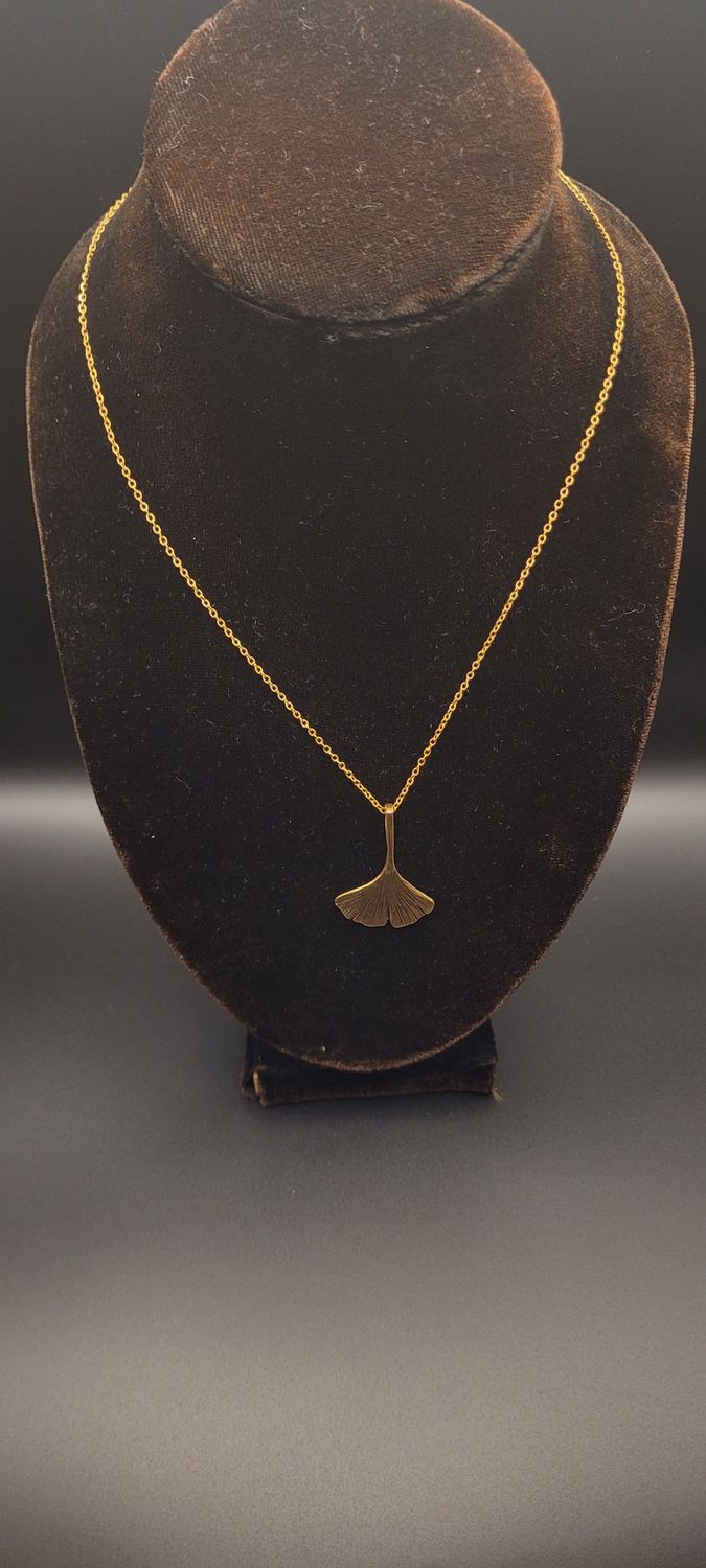 A a gold metallic ginkgo leaf pendant on a gold chain and a display stand.