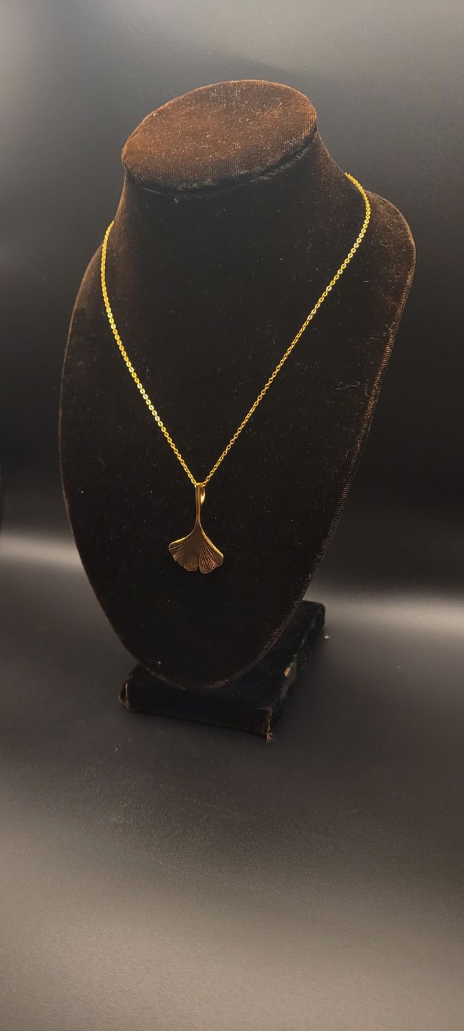 A a gold metallic ginkgo leaf pendant on a gold chain and a display stand.