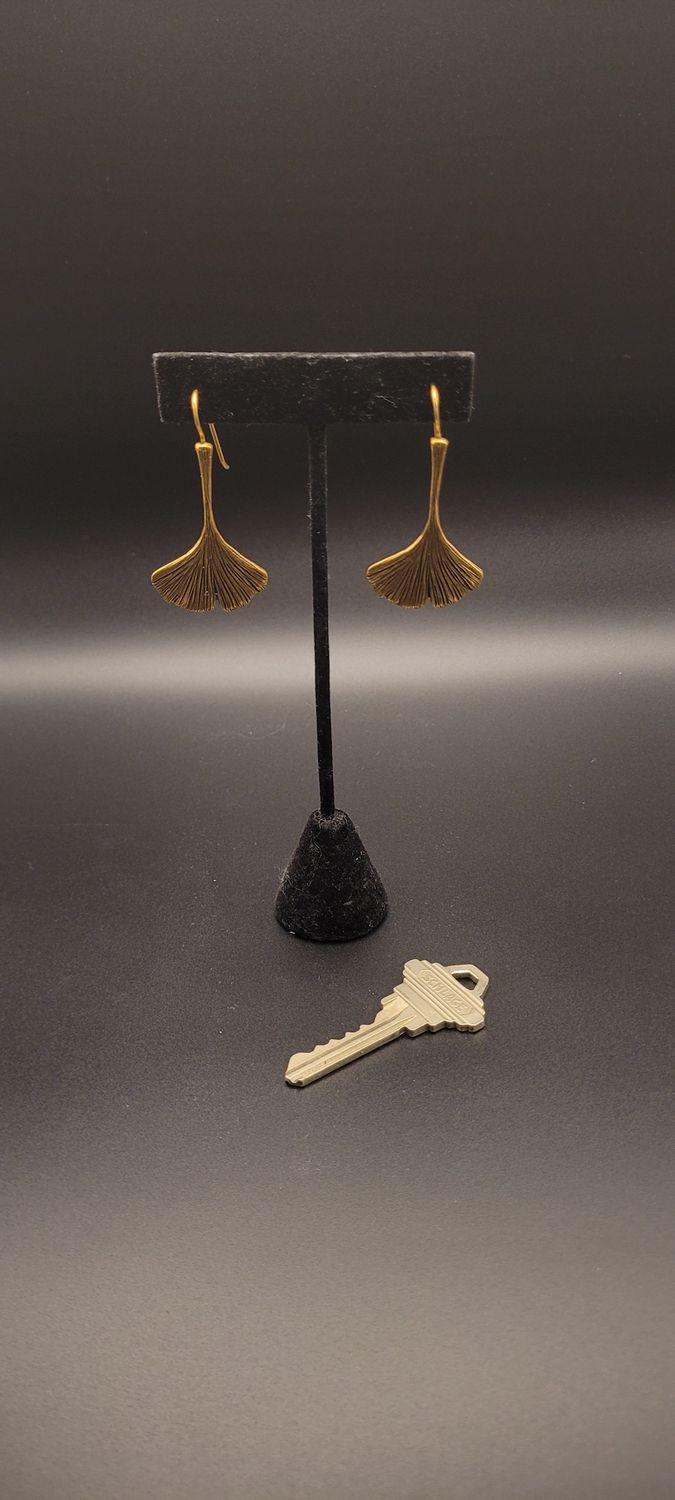 Two gold-metallic earrings featuring a ginkgo leaf on a black stand.  There is a housekey for scale.