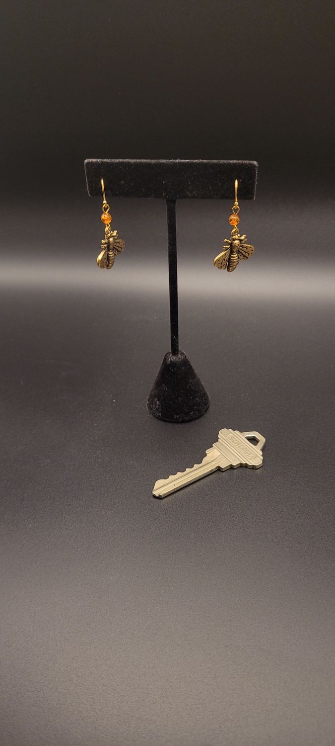 Two earrings, each with an orange amber bead and then a metallic bee beneath them.  There is a key for scale as well.