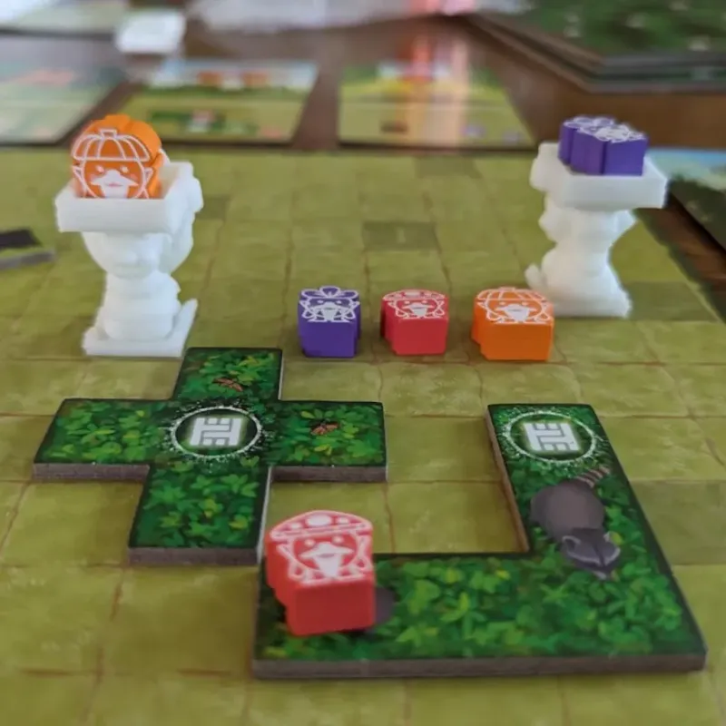 Hedge Mage game board with green tiles, white pedestals, and colorful player tokens, featuring orange, purple, and red pieces on display.