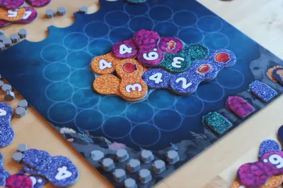 Lumicora board game pieces on a blue game board, featuring colorful coral tiles with numbers, for 2-4 players aged 10 and up.