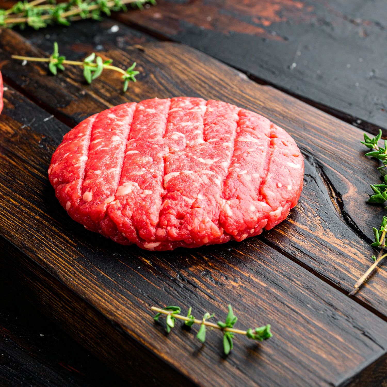 Raw angus ground beef patty with grill marks sits on a wooden cutting board, surrounded by sprigs of fresh thyme.