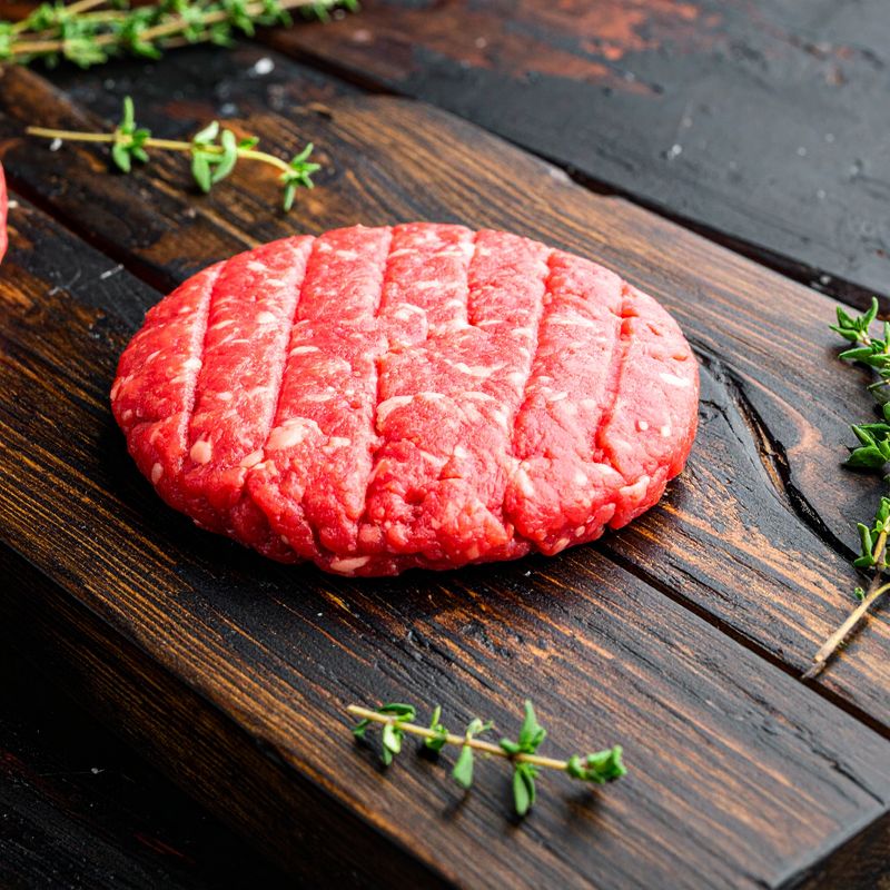 Raw angus ground beef patty with grill marks sits on a wooden cutting board, surrounded by sprigs of fresh thyme.