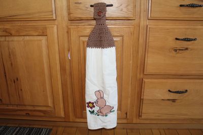 Hand painted bunny kitchen towel with flower and brown crochet topper hanging on cabinet handle.