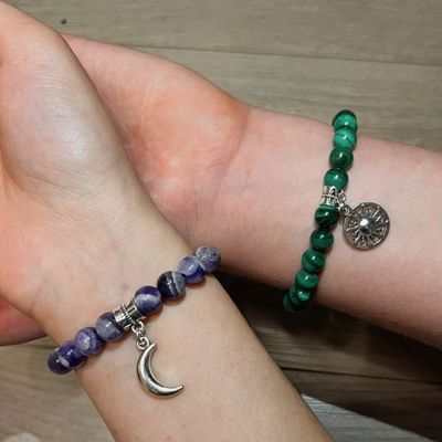 Two crossed forearms with bracelets. One is amethyst, violet. Second one is malachite, green. Metal charms - sun and moon.