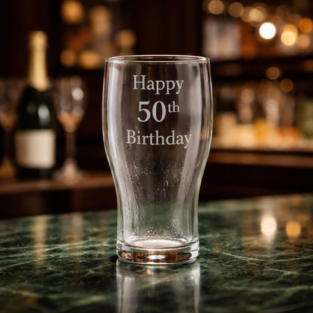 Personalised engraved clear glass pint glass sitting on a green marble bar top, engraved with the text "Happy 50th Birthday".