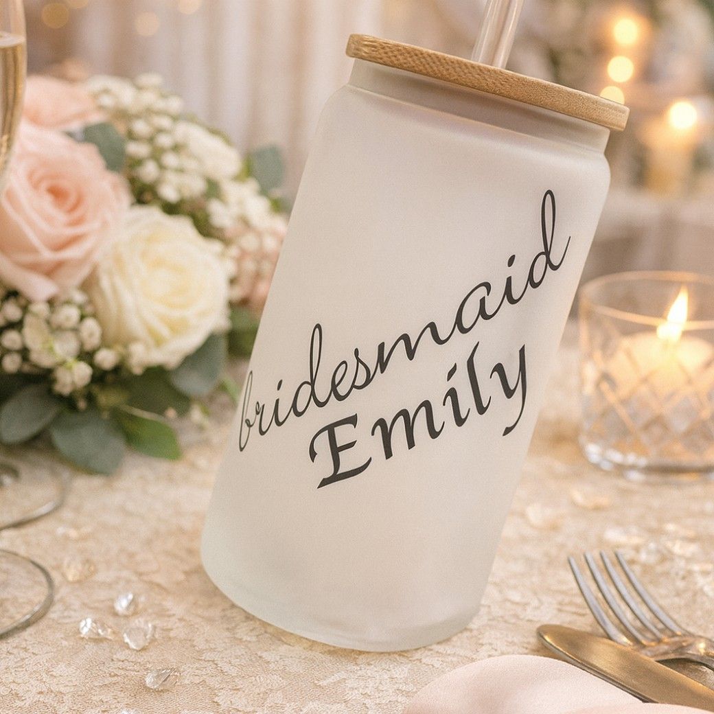 A frosted tumbler with bamboo lid and straw reads “bridesmaid Emily” in script, placed on a lace-covered wedding table with f