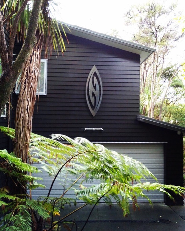 Grounded Art NZ Corten Steel Wall Art Shield in Fish Hook Design on the exterior wall of a NZ home. Native NZ garden setting.