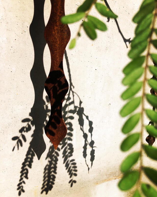 Corten Kowhai pod by Grounded Art NZ. Hanging infront of a natural stone wall casting shadows of the pods and Kowhai leaves. 