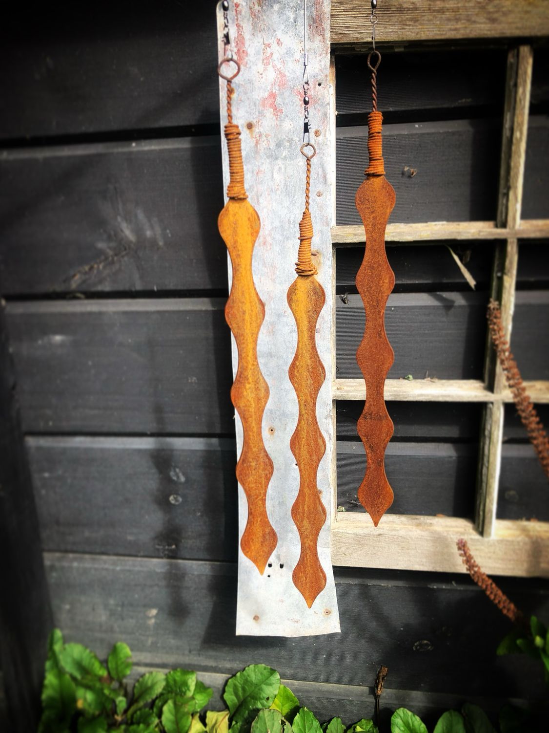 3 Corten Kowhai pods by Grounded Art NZ. Hanging in front of a dark wall and old window the Corten stands out in contrast.