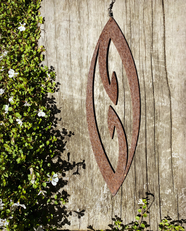 Rusted Corten Fish Hook (mini) Spinning Art with a weathered steel finish in a landscaped garden setting.