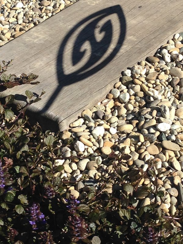 Shadow cast by Fish Hook Aluminium Garden Art Spear by Grounded Art NZ falling across a wooden paving step and pebbles.