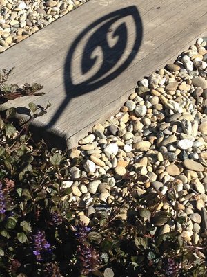 Shadow cast by Fish Hook Aluminium Garden Art Spear by Grounded Art NZ falling across a wooden paving step and pebbles.