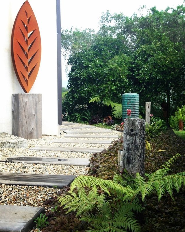 3D Corten Leaf Sculpture by Grounded Art NZ in a wooden base, natural stone and wooden path in a lush green native garden. 