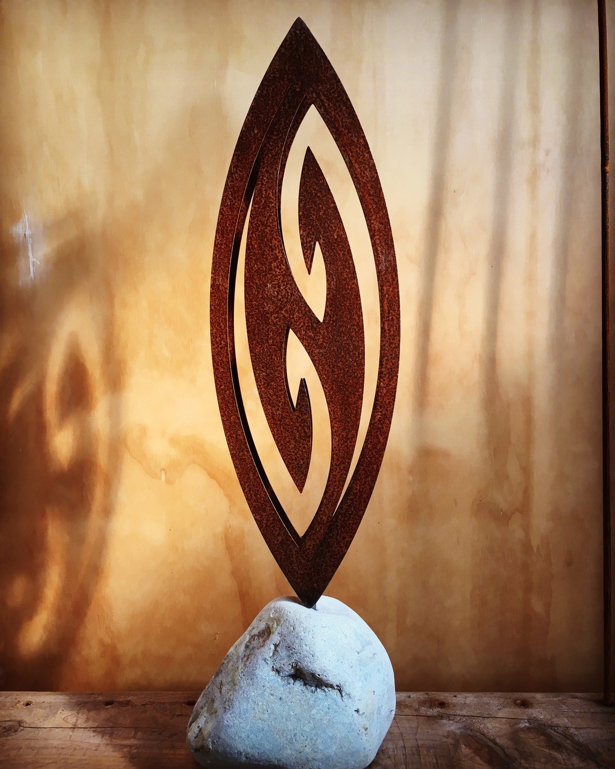 3D Corten Fish Hook in a stone base. shadows cast on wall behind in a warm, natural looking gallery setting.