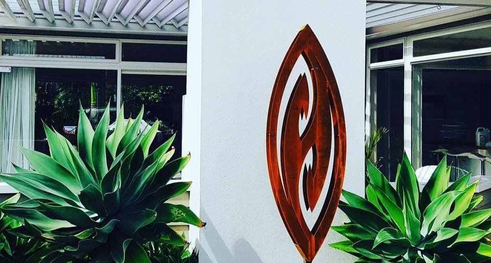 Rusty 3D Corten Fish Hook Sculpture by Grounded Art NZ in a white courtyard surrounded by lush tropical plantings.