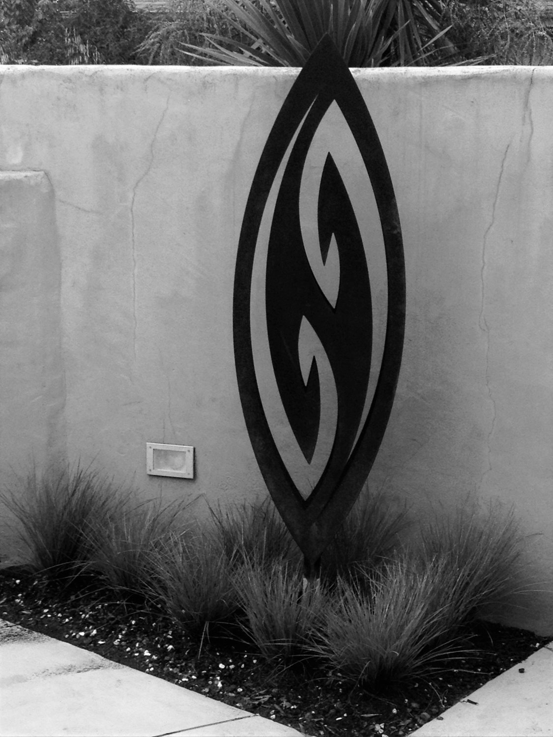 Corten Fish Hook by Grounded Art NZ in black and white. Courtyard setting with grasses and lighting beneath the sculpture. 