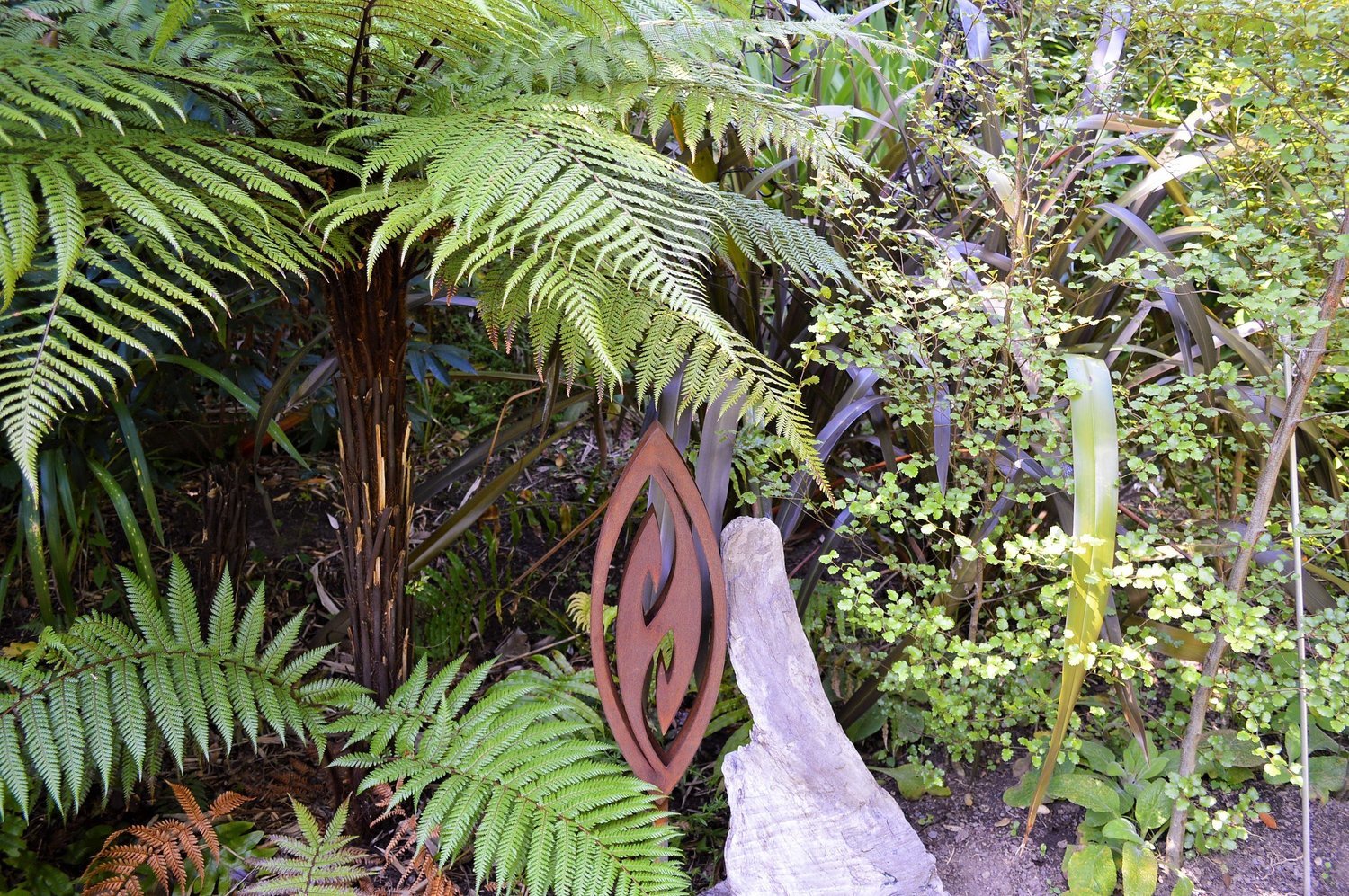 A 600mm 3D Fish Hook in rusted Corten Steel displayed in an established Native garden. Garden Art by Grounded Art NZ.