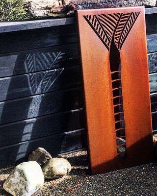 Corten Nikau panel by Grounded Art NZ leant up against a black wall, shadows are created behind the panel. Bright Patina.