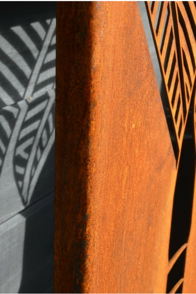 Corten Nikau panel by Grounded Art NZ leant up against a black wall, shadows are created behind the panel. Bright Patina.