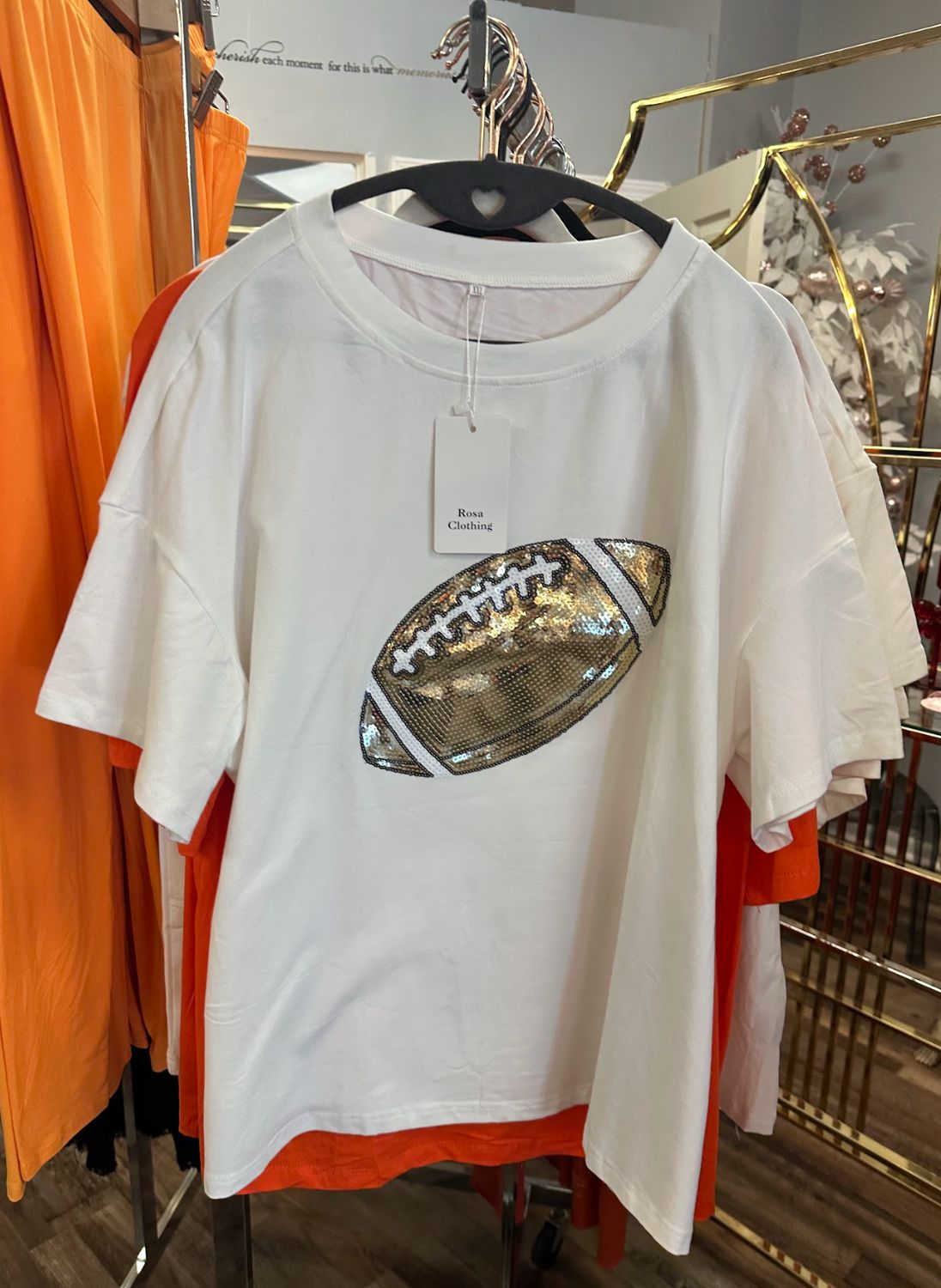 Rosa Clothing Football  T-shirt in White & Shimmering gold hanging on a rack in Merle Norman Elizabethton. 