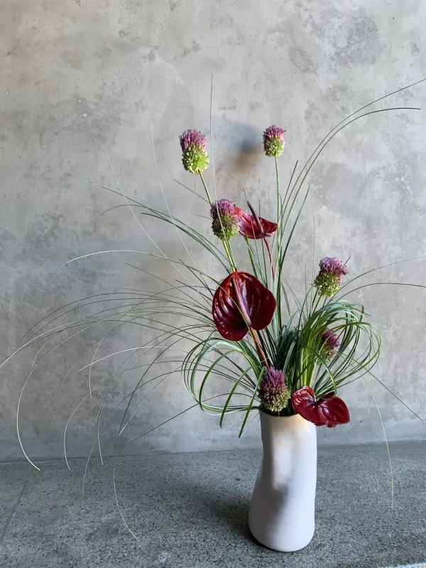 Vogue arrangement of deep red anthuriums, purple allium blooms, and dramatic ornamental grasses in a white ceramic vase