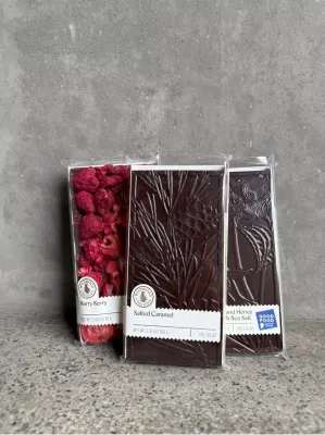 Three artisan dark chocolate bars in clear packaging featuring berry, salted caramel, and floral embossed designs