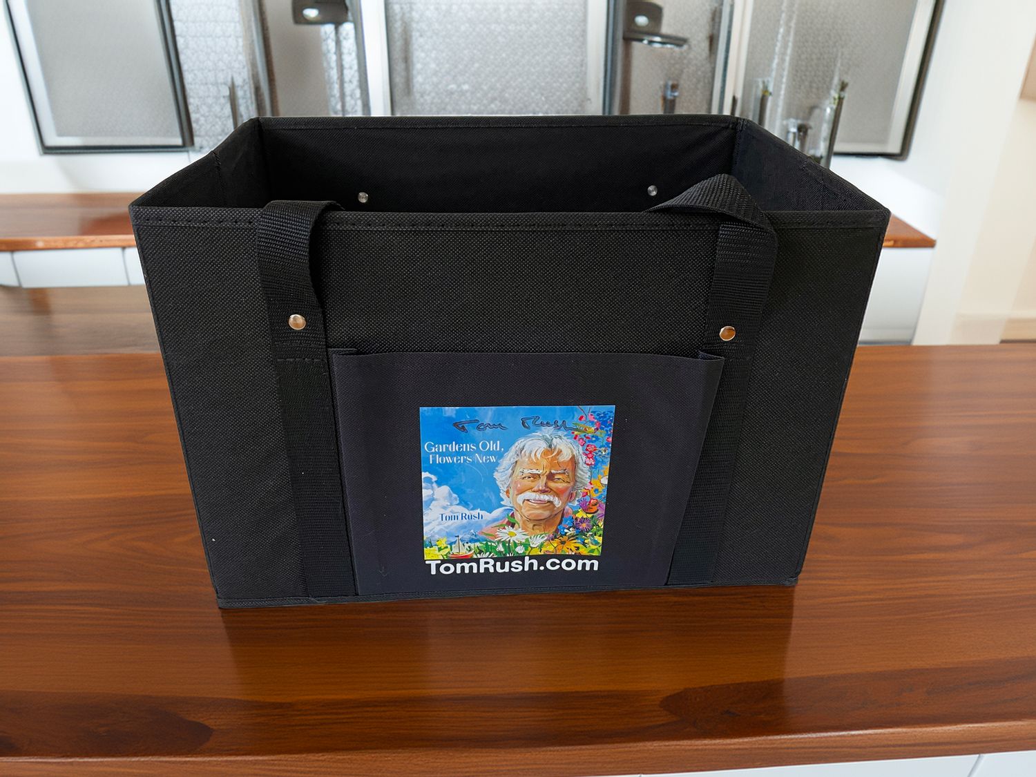 A black rectangular tote bag sitting on a tabletop.  An image on the front features an drawn image of Tom Rush, the words, TomRush.com, and his signature