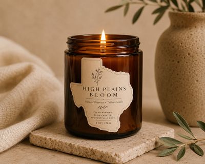 Lit amber glass candle labeled “High Plains Bloom” centered on a stone coaster with clean linens and minimal esthetic. 