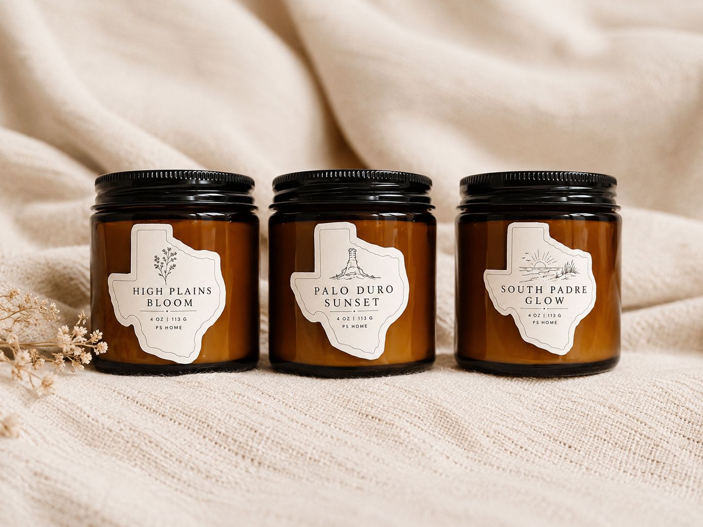Three amber glass candle jars from the PS Home signature collection are arranged side by side on a soft cream knit blanket. 