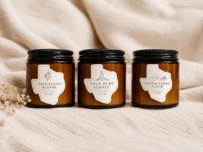 Three amber glass candle jars from the PS Home signature collection are arranged side by side on a soft cream knit blanket.