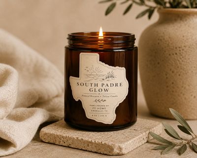 Lit amber glass candle labeled “South Padre Glow” set against a stone coaster and clean linens evoking elevated and natural 