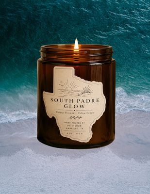 Lit amber glass candle labeled “South Padre Glow” set against an aerial ocean shoreline, with turquoise waves and white sand