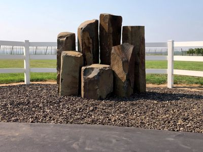 Grouping of six basalt columns set in a black crushed rock bed at a vineyard entrance, white rail fence behind.