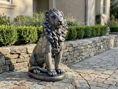 Dark cast concrete lion statue seated on a paver landing beside a stacked stone wall and clipped boxwood hedge.