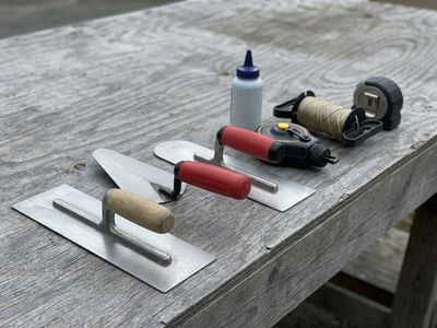Concrete finishing trowels, a chalk line, mason line and a tape measure laid out on a weathered wood bench.