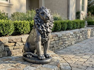 Dark cast concrete lion statue seated on a paver landing beside a stacked stone wall and clipped boxwood hedge.