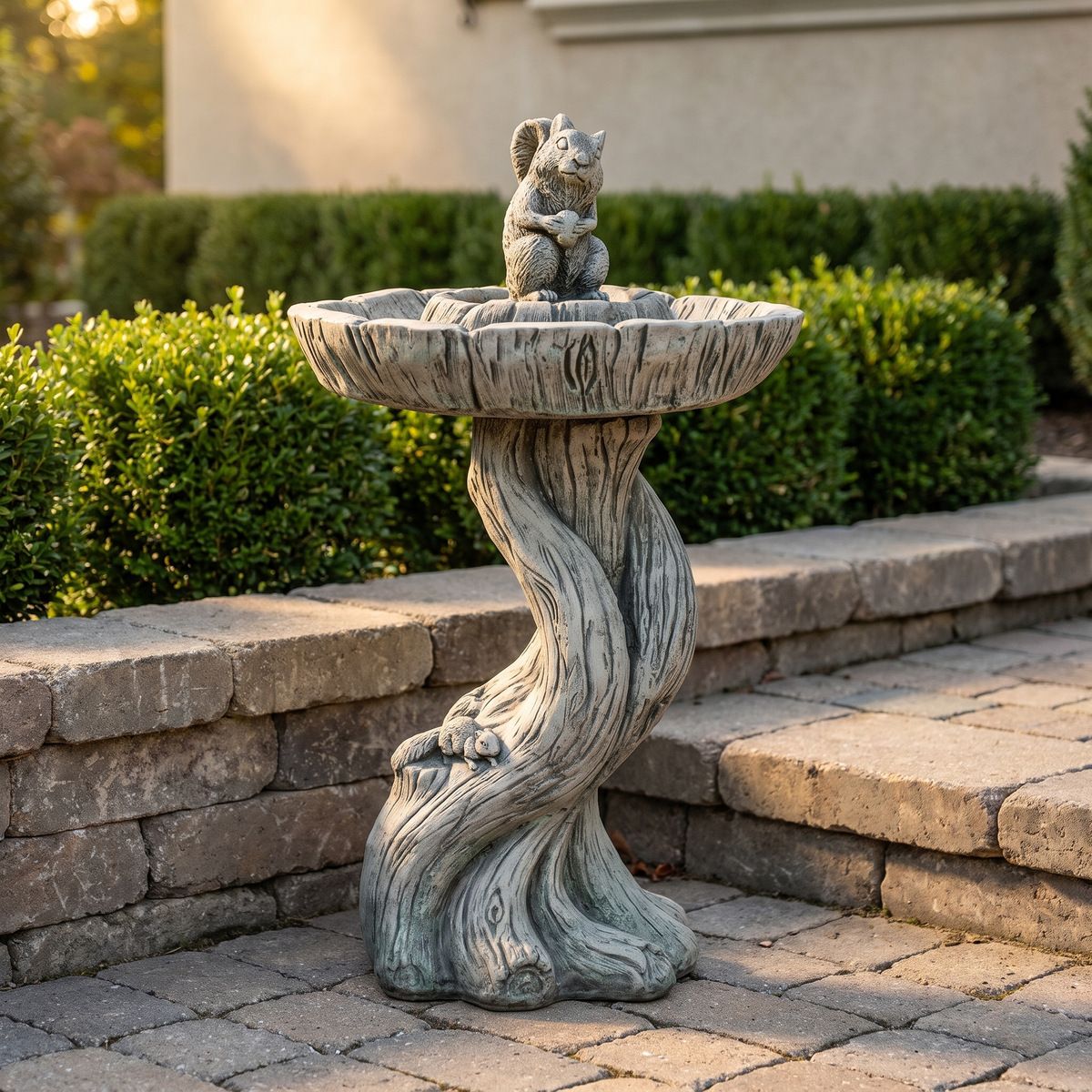 Concrete squirrel bird bath on a woodgrain pedestal, set on pavers beside a stacked stone wall.