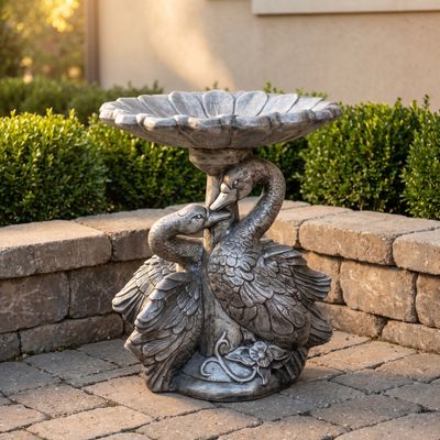 Double swan concrete bird bath with a scalloped bowl, on a paver landing beside a stacked stone wall.