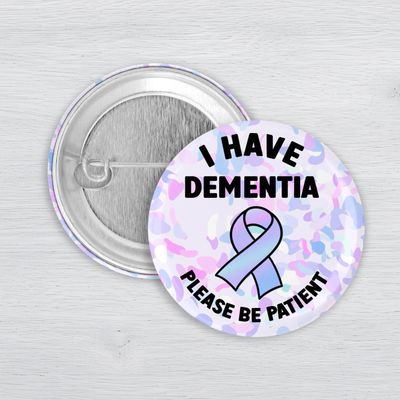 Dementia awareness pinback button with pastel ribbon and “Please Be Patient” message on white background.