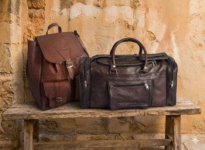 Ein handgefertigter Leder-Rucksack und eine dunkle Ziegenleder-Reisetasche der Traveler Pro Serie auf einer rustikalen Holzba