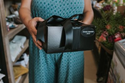 Woman holding a black Urban Market 1919 gift box with a ribbon, representing locally inspired gifts.
