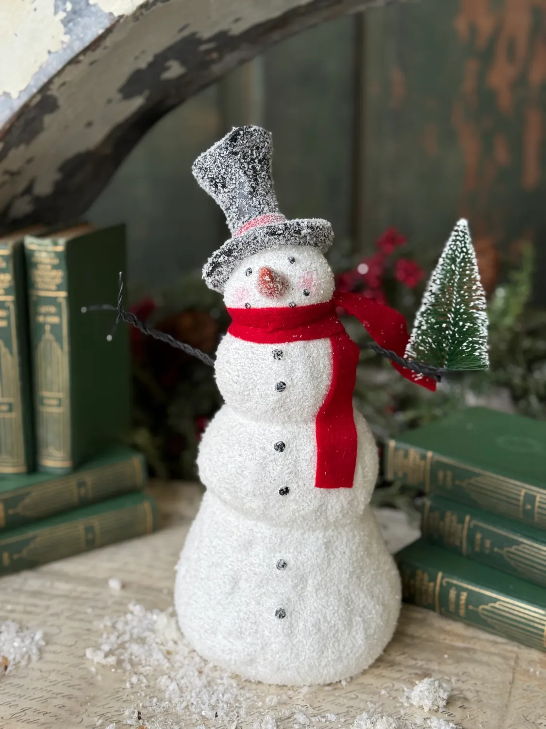 Blizzard Boy Snowman I 11.5", a festive snowman figurine with red scarf and hat, standing on a surface surrounded by books and snowflakes.