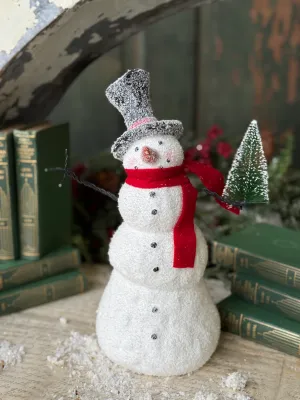 Blizzard Boy Snowman I 11.5", a festive snowman figurine with red scarf and hat, standing on a surface surrounded by books and snowflakes.