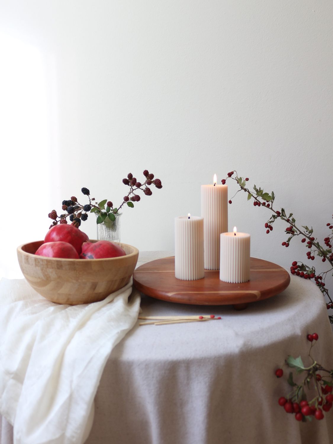 Trio di candele a colonna a costine in tre misure accese su una tavola con frutta e bacche