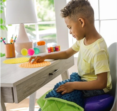 A boy sits doing classwork with a weighted blanket on his lap. He is sitting on a textured wiggle pillow.